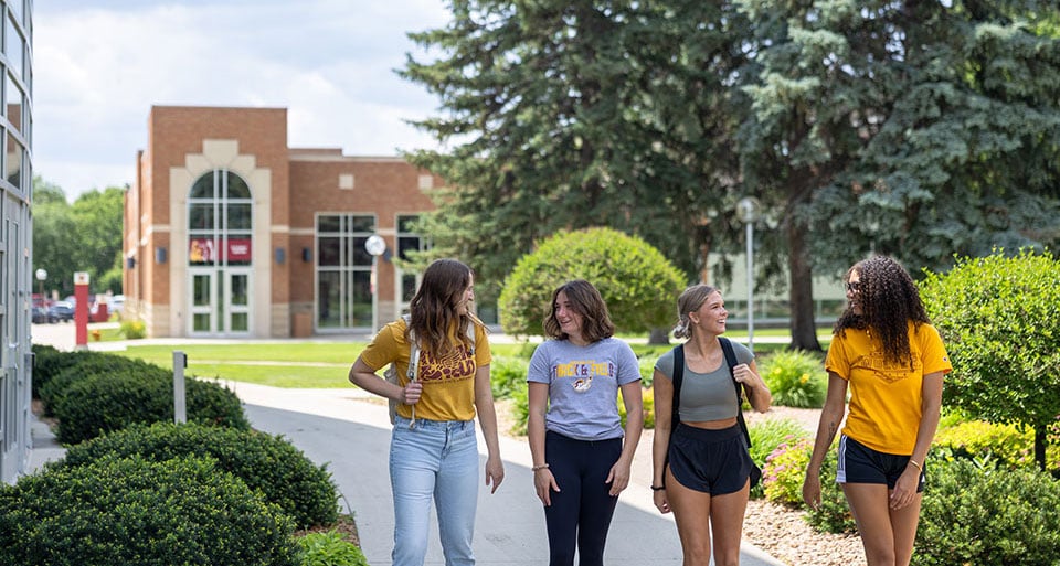 Four students walking on campus sidewalk, smiling and talking